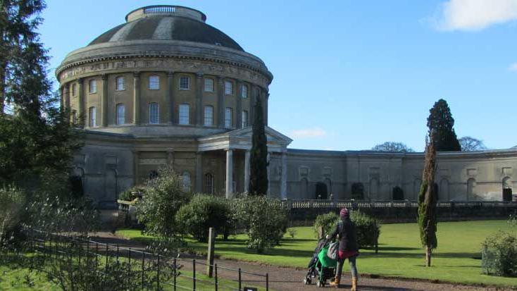 The rear of Ickworth House - a domes central rotunda with wings on either side. A woman walks with a pushchair towards the house along a gravel path with lawns either side.