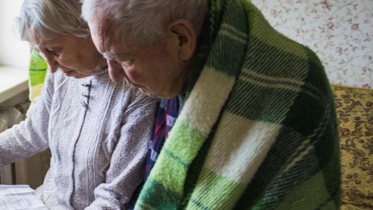 An old woman with a heating bill by a radiator and an old man in a green blanket