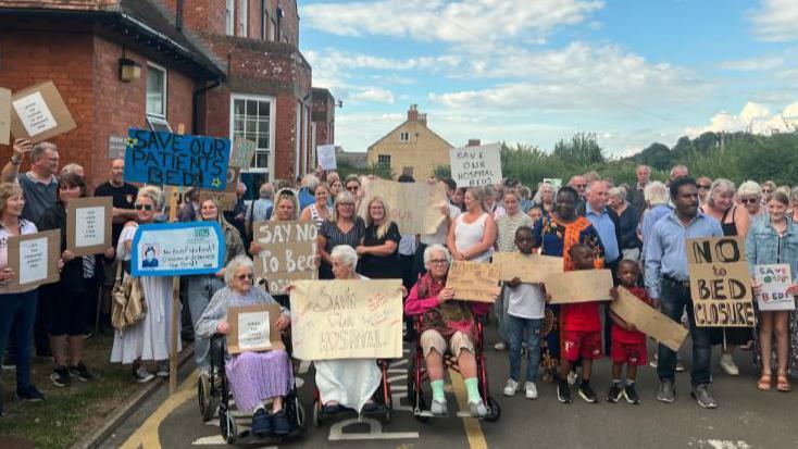 A crowd of people stand outside a red brick building carrying signs saying "Save Crewkerne Hospital", "say no to bed closure" and "help us fight".