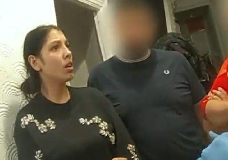 A woman with dark hair in a low bun is standing in her hallway, wearing a black jumper with white flowers on. She looks shocked.