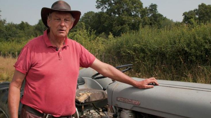 A farmer in a red shirt with a hat on stands with one hand on a tractor as he speaks to the BBC