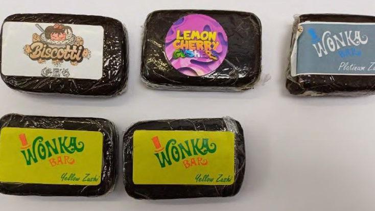 Five brick-sized packages of cannabis in black bags. The packages have colourful stickers on them. The bottom two read 'Wonka Bar' on them while the top read Biscotti, Lemon Cherry Crusher and Wonka Platinum.