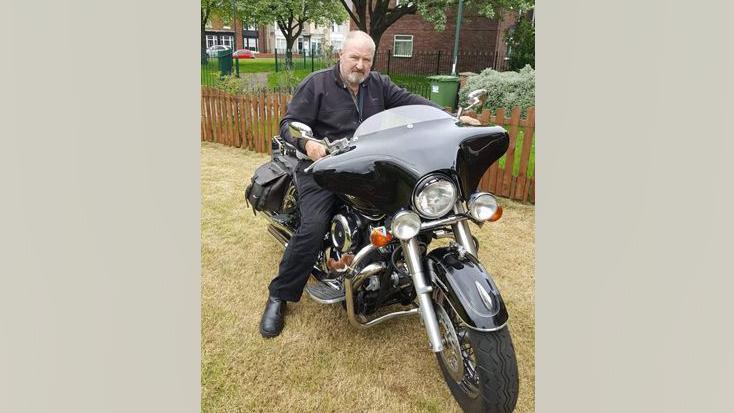 John Dingwall sitting on a black motorbike. He is wearing a dark shirt. He has short grey hair and a grey shortly cropped beard.