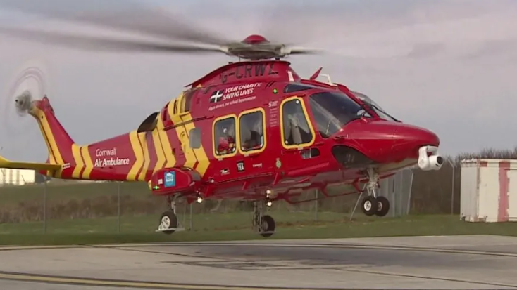 A red and yellow helicopter facing towards the right hand side of the image starts to take off from a tarmac helipad. The propeller blades are spinning. 