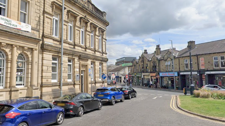 Thornton Square, on Briggate in Brighouse. Cars are parked in on-street bays on the left with the high street in the background. 