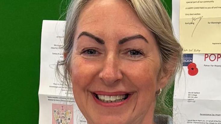 A smiling Vicky Pudney looking straight at the camera. She is standing in front of a green noticeboard with several letters pinned to it.