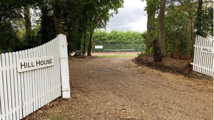 Hill House sign in black lettering on white picket style fence either side of a gravel driveway between trees with what appears to be a clay surface tennis court in the background 