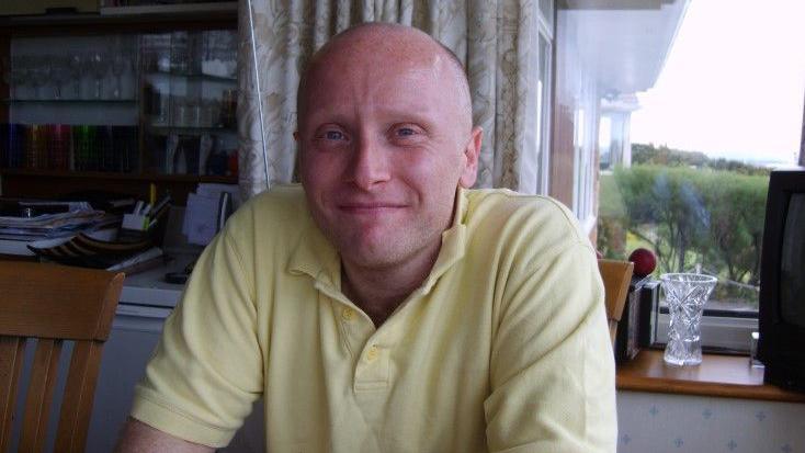 Dan Kay, who is bald and wearing a yellow polo-neck t-shirt, smiles at the camera while sat in a kitchen