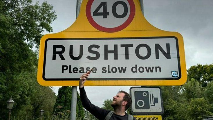 a man looks frustrated holding a mobile phone in the air in front of a road sign reading "Rushton, please slow down