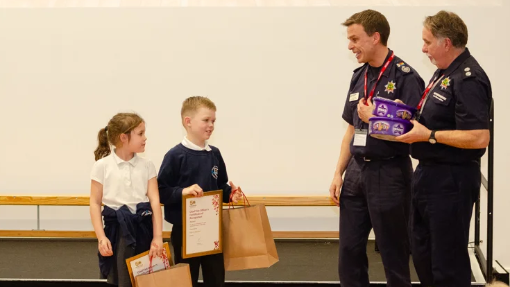 Harry and Grace holding certificates and paper bags. There are two fire service employees wearing navy uniform and holding two boxes of Heroes.