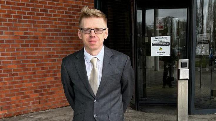 A picture of Councillor John Baker, stood outside Central Bedfordshire councils headquarters in Chicksands. He is wearing a dark grey suit with a white shirt and silver tie and is wearing glasses.