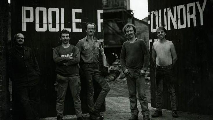 Black and white photo of five men posing in front of the open gates. The words 'Poole Foundry' are written above their heads across the gates.