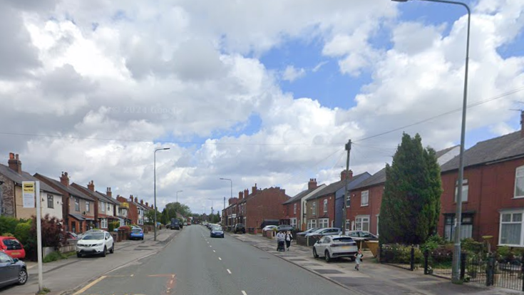 A car drives down Wigan Road, with people walking along the pavement on either side