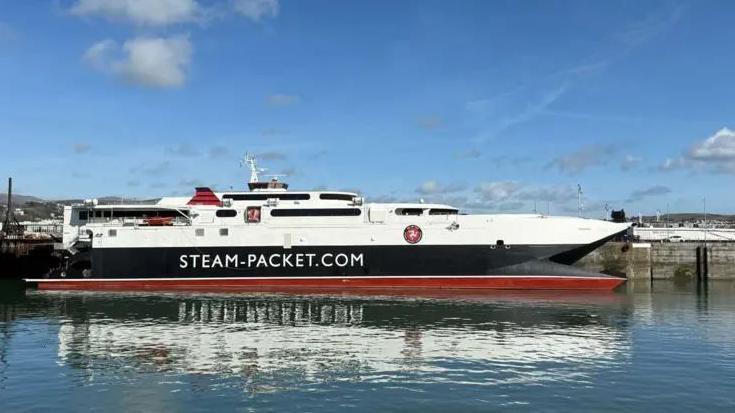 The Manannan ferry in Douglas Harbour. It is white, red and black and has STEAM-PACKET.COM on the side. The sky above is blue.