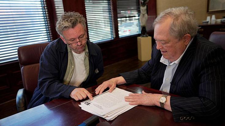 A meeting takes place around a polished wooden boardroom table, where Sheen and Stewart sit side by side reviewing a printed report. Stewart, who has grey hair and is wearing a pinstripe suit points to text on the top page while Sheen rests a hand on the document. Sheen is wearing a navy overshirt over a green shirt and white T-shirt. Large windows with partially closed blinds line the wall behind them, and office furnishings and artwork can be seen in the background.