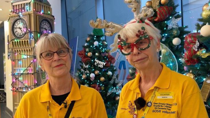 Two Great Ormond Street Hospital volunteers in yellow uniforms stand in a festive hospital lobby decorated with Christmas trees and a model of Big Ben, one wearing reindeer antlers and holiday-themed glasses.