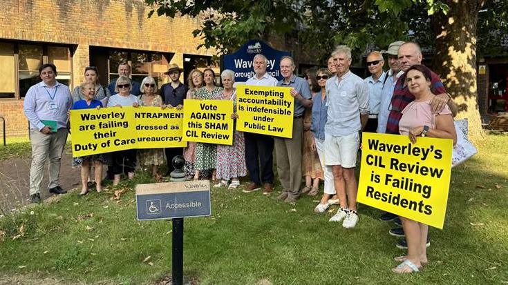 A group of people standing on a grass patch with a sign behind them that says "Waverley Borough Council". Some of them are holding yellow signs criticising the council.