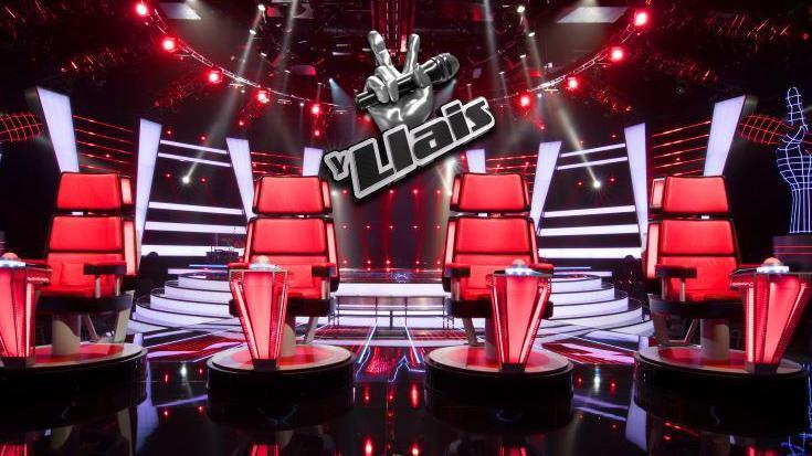 Four red chairs facing the camera. Behind them is a stage and a sign reading Y Llais