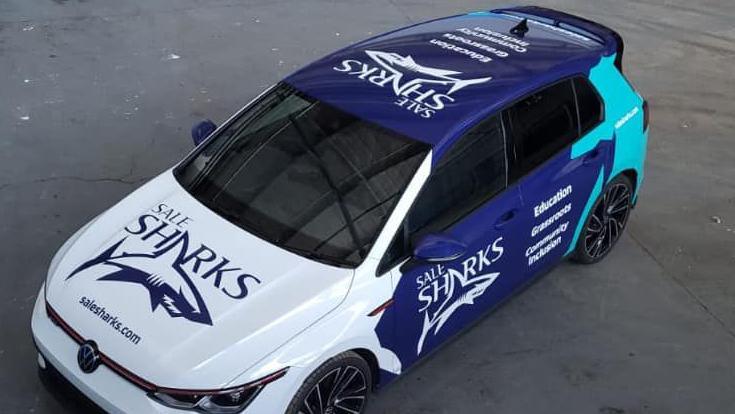 A VW Golf Club Sport car, with white, blue and teal painting on it, with Sale Sharks and a shark logo painted on it. 