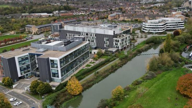 Aerial shot of the UON Waterside Campus, showing river in the centre with marina to the right, and modern grey and white university buildings to the left.  There are houses in the background.