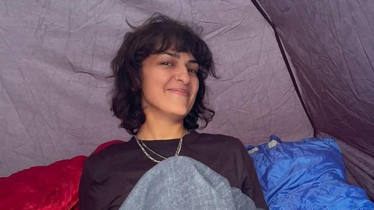 Henrietta, a woman with short brown hair and a fringe, smiles. She is sitting down in a tent and is wearing a long-sleeved black top and two necklaces around her neck.