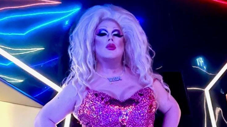 Vida LaFierce stands in front of a bright background, she has a large blonde hairdo and striking makeup with large lashes. She wears a necklace that reads Vida and a pink sparkly strappy top.