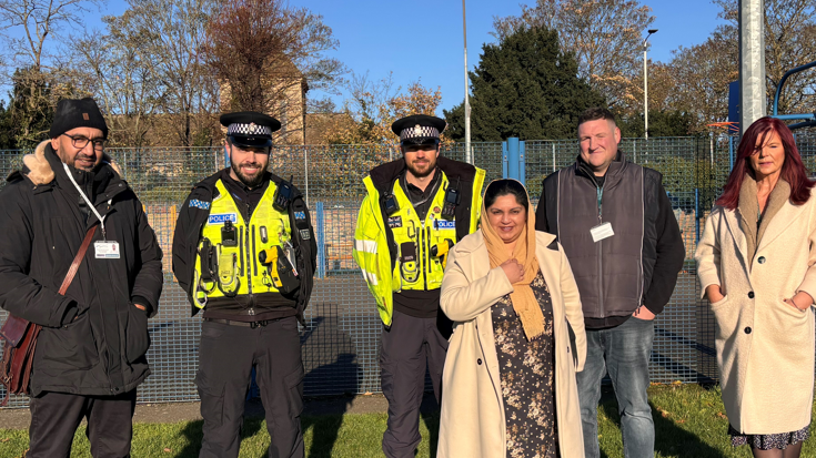 Two police officers wearing high visibility coats and vests are stood in a park with two men and two women. 