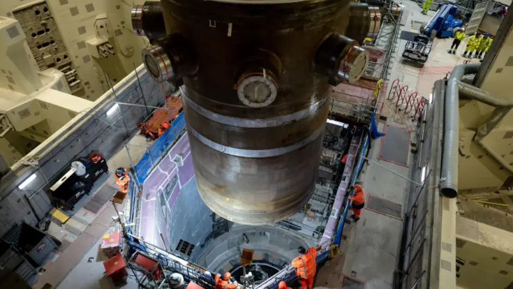 A massive nuclear reactor being lowered into Hinkley Point C chamber. There are construction workers watching.