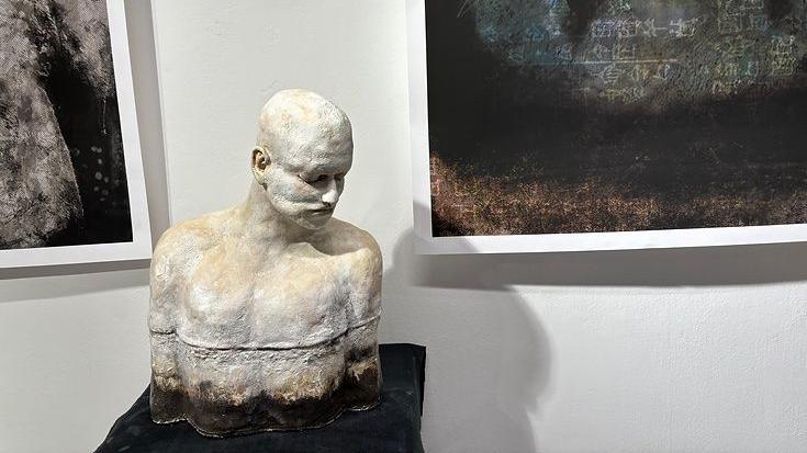 A clay sculpture of a man looking down and looking sad. It is sitting on a black shelf and behind it you cam see parts of paintings 