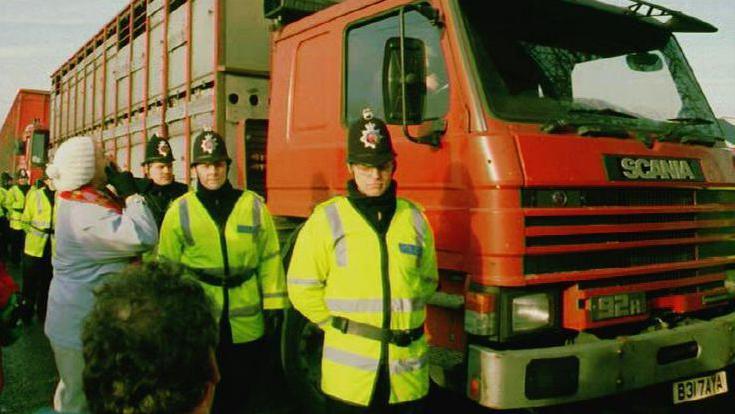 A row of police officers standing next to a red lorry. A protester is standing next to one of the police officers.