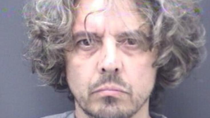 A police custody photo of Ben Atkins, he has curly grey hair over his ears and is unshaven. He is wearing a grey top and standing against a dark grey wall