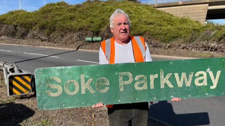 Mark wearing a white T-shirt and an orange vest holding a long green Soke Parkway sign with both his hands - standing on the slip road.