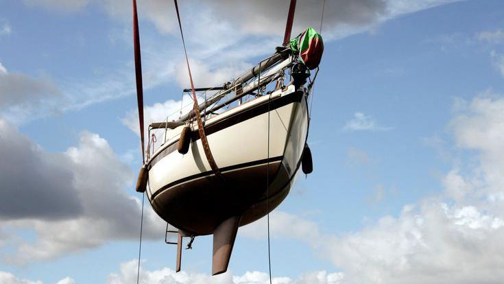 A small white sailboat is suspended by straps from a shipyard crane for seasonal repairs.