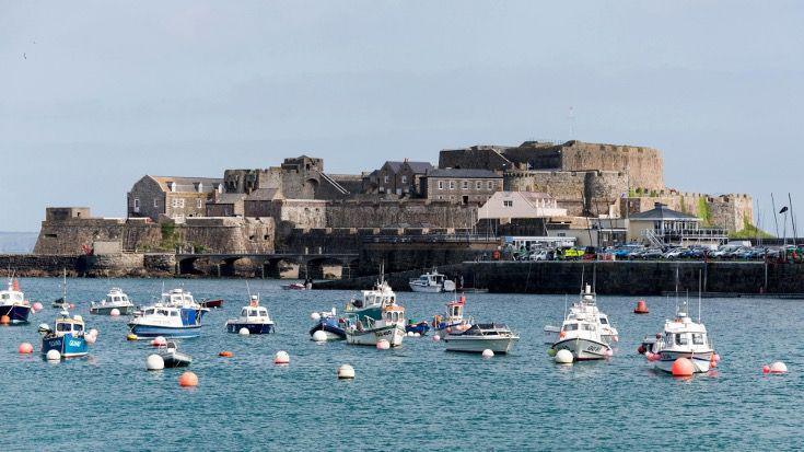 Boats are pictured within a patch of water. There is buildings in the background of the shot with a car park next to it. 