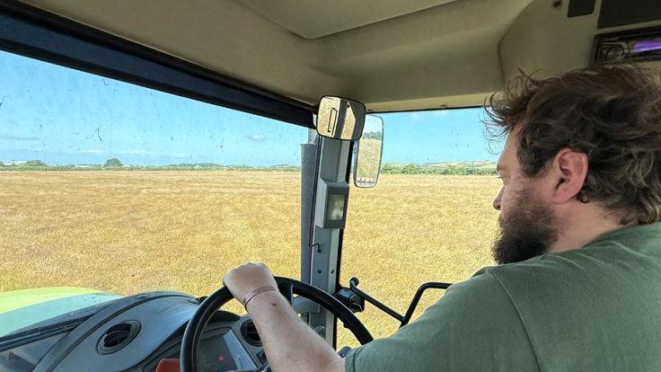 The view from the passenger seat of a tractor. Sam Morrey wears a green T-shirt, he drives a combine harvester. Through the windows you can see large fields of yellow crops.
