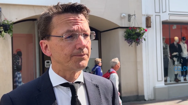 Deputy Kirsten Morel, a man with dark brown hair and rimless glasses, wearing a dark suit and tie and a white shirt. He is looking to the right of the screen while standing on a pedestrian street in St Helier, Jersey. Behind his head, baskets of pink and purple flowers are hanging from the wall of a building.