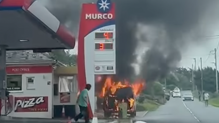 A car is on fire next to the petrol station, it has dark plumes of smoke coming from it and orange flames. A person is seen looking on. There is a sign that says MURCO