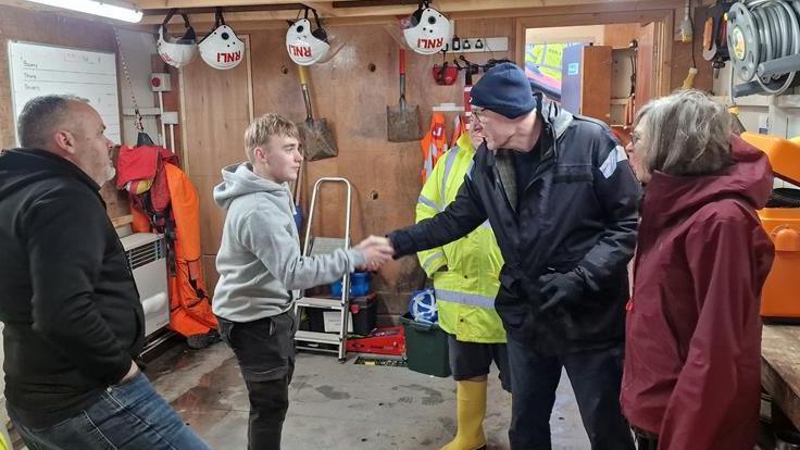 Rob Beckley is saking the hand of a young man. They are standing with three other people in a shed filled with rescue equipment.