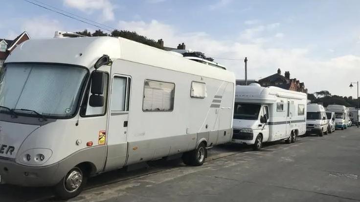 A row of light coloured campervans, parked up bumper to bumper on a road. Above can be glimpsed roofs of buildings. In front is a wide tarmacked road or pavement.