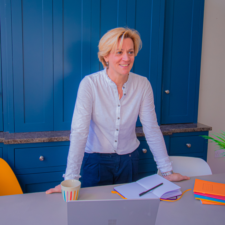 Picture of Anna Doneghey wearing a white shirt and standing at a desk with an open notebook, mug and laptop on it. 