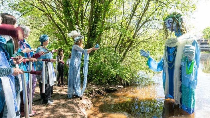 A large blue river goddess puppet stands in shallow river water, while people dressed in blue clothes stand on the bank. One of them is standing at the water's edge. She wearing a long blue dress and a bird headdress, and reaching out towards the puppet while holding a blue object in her hands