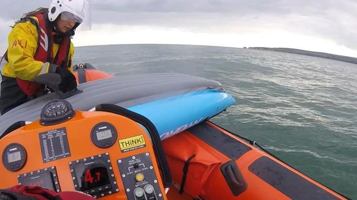 View from on board the orange RHIB lifeboat with a lifeboat crewmember in yellow and red securing the blue and grey kayak