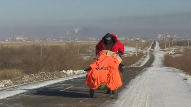 Karl Bushby pushes a buggy containing his belongings along a snow-covered road, which passes through scrubland to either side. He is wearing a balaclava, sunglasses and a red coat. Behind him, a city can be seen in the distance.