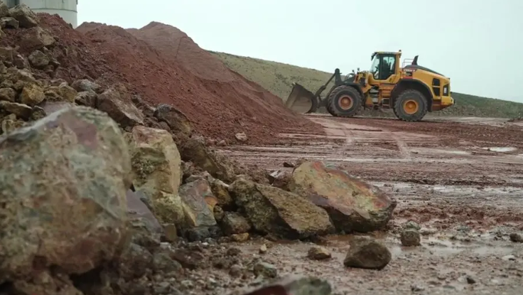 A yellow digger drives through a quarry. In the foreground there are large lumps of rock and piles of red earth. 