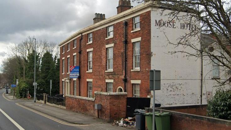 A building with red brick and traditional feature windows on the front and a white painted side, with painted letters reading Moose Hotel. Rubbish is strewn on the street next to several wheelie bins. 