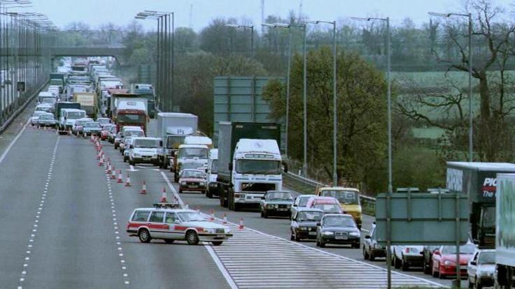 A vintage looking police car protecting a line of cones diverting drivers off the motorway at a junction slip road, with heavy congestion on the approach