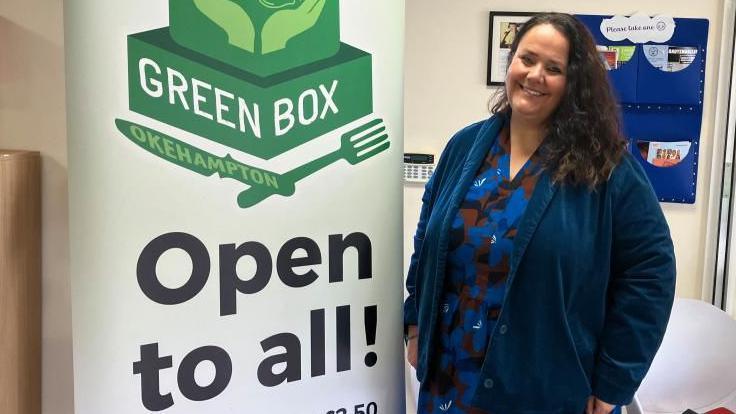 Melissa Trudgill, community engagement officer for Dartmoor Multi Academy Trust is wearing a dark blue patterned dress in front of a sign which reads: Green Box, Okehampton, Open to all!