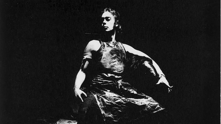 A man dressed in a dark corset on stage. The stage and everything else is entirely dark - only he is illuminated.