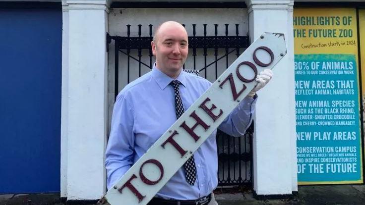 Man in shirt and tie holding a big white sigh that reads "To the Zoo"