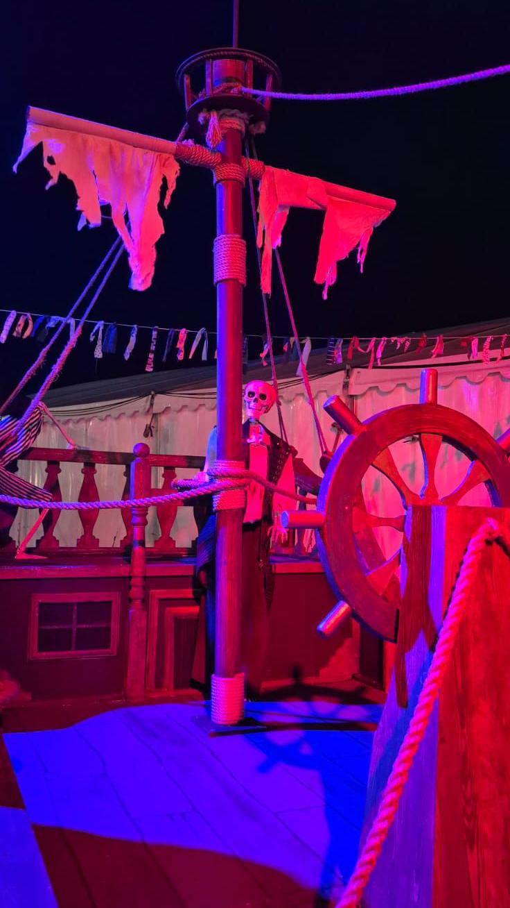 A skeleton stands on the deck of a wooden pirate ship, with red lighting.